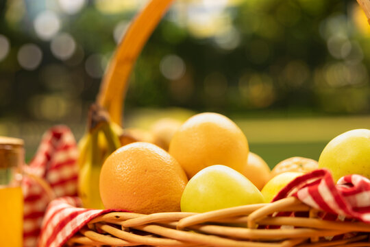 Fresh oranges and apples fill a wicker picnic basket lined with checkered cloth, illuminated by warm sunlight in a public park during a relaxed outdoor picnic.