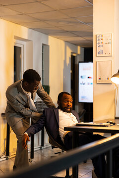 Black office worker playfully startles a sleepy colleague who has fallen asleep during late-night overtime, revealing the mental exhaustion and workplace pressure caused by ambitious deadlines.