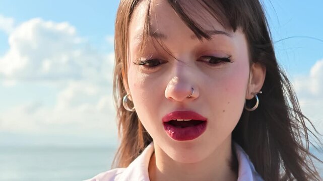 Shocked young woman with wide open mouth and surprised expression by the sea, wind blowing her hair, amazed reaction, astonishment, disbelief and wow emotion concept on a sunny day with blue sky 