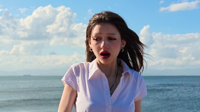 Shocked young woman with wide open mouth and surprised expression by the sea, wind blowing her hair, amazed reaction, astonishment, disbelief and wow emotion concept on a sunny day with blue sky 