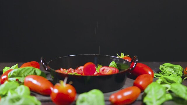 Tomatoes and basil fall into a black cooking pot, surrounded by ingredients on a dark background