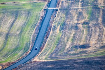 Car driving on narrow paved road through textured agricultural fields. The road divides patterned green and brown hillsides, capturing a theme of road trips, transit, and isolation in rural landscape © aquamarine4