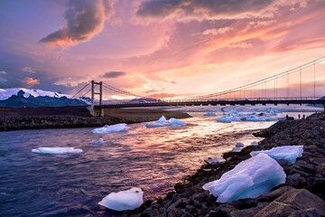 Suspension bridge over Glacier lagoon with icebergs at sunset. Brilliant pink sunset clouds illuminate the sky above the water and distant snow-capped mountains in an Arctic landscape in Iceland.   © aquamarine4