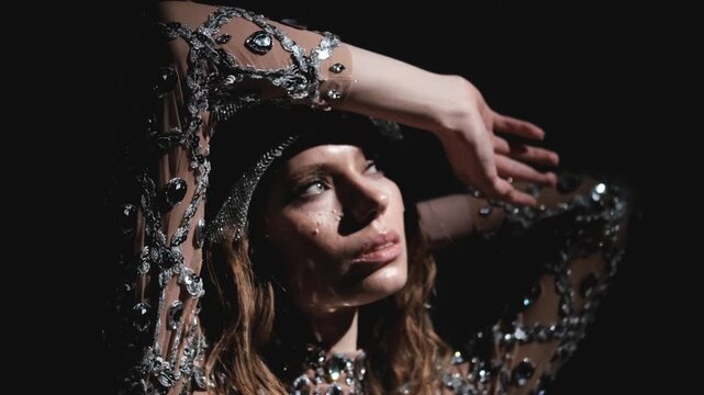 A woman in a sparkling outfit posing with her hand on her head, in dark background, with sparkling dress, hand, from a close-up perspective, with brown, silver accents.