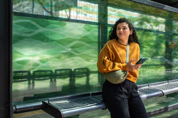 Smiling woman tourist wearing a fanny pack waits at a bus stop, holding a phone and looking into the distance  a young traveler using public transport to explore the city © Victor Borges