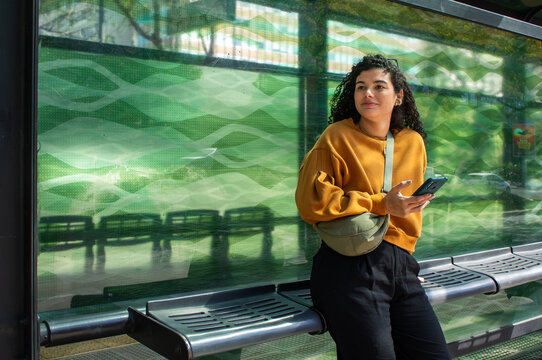 Smiling woman tourist wearing a fanny pack waits at a bus stop, holding a phone and looking into the distance; a young traveler using public transport to explore the city