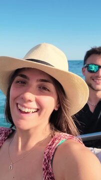 Couple takes a selfie while riding a boat cruise across the ocean. Young woman smiles, talks and turns toward the sea as her partner enjoys the sunny journey, capturing a joyful summer vacation.