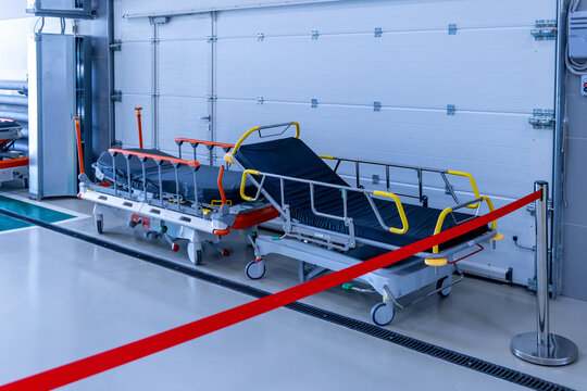 Two empty hospital stretchers with raised side rails stand by a closed garage gate in the ambulance bay. A red belt barrier fences off the area. The trolleys are ready for incoming patients