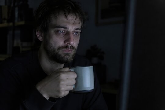 Exhausted young man holding coffee cup while working late night on computer in dark room, feeling tired and stressed, insomnia concept