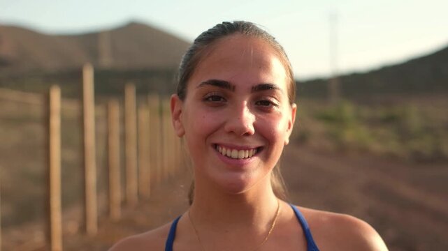 Woman enjoys evening energy after a rural workout, smiling at camera and tucking hair behind her ear. Warm sunset light and open hills convey active lifestyle, health, wellness and optimism.