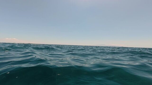 Ocean waves ripple across calm blue water as the water-level POV camera gently bobs toward a distant horizon. Clear sky and sunlight create a peaceful sea background for travel, summer and wellness.