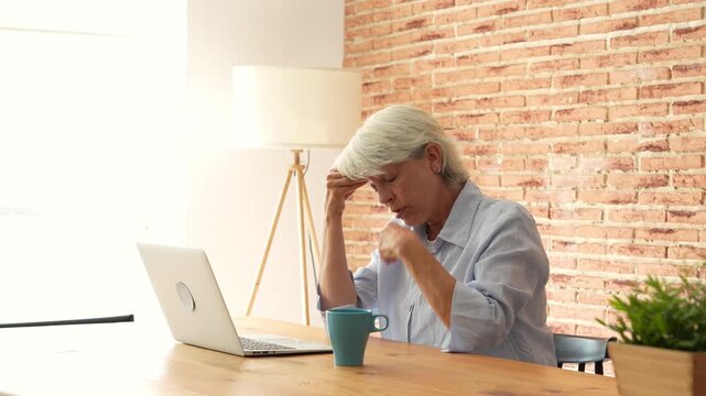 Senior woman massages her temples, leans back and runs her hands through white hair as a headache brings stress and fatigue while working on a laptop at a wooden desk in her exposed brick home office.