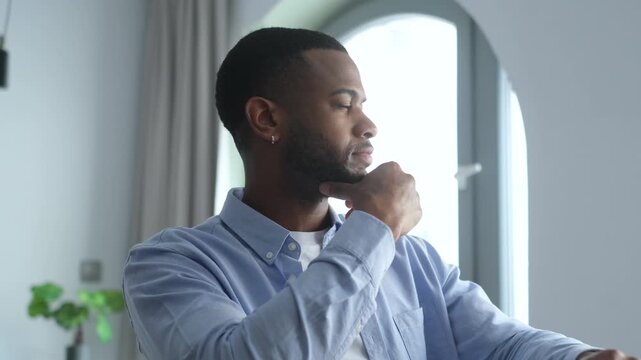 Black man pauses from remote work at home, resting his chin on his hand while looking out the window. Thoughtful freelancer sits in a bright home office, considering a work problem with focused calm.