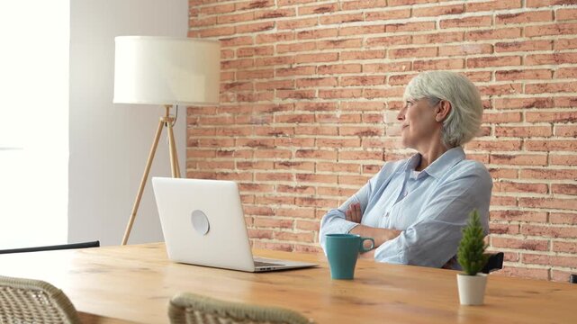 Senior woman types on a laptop at home, then pauses, looks away and smiles while reflecting at her desk. The clip conveys remote work, aging, technology, workplace stress and work-life balance.