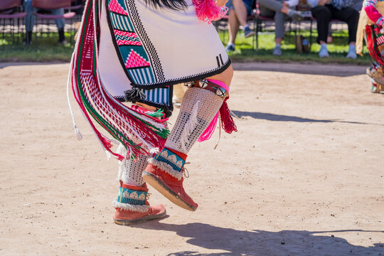 Detailed view of colorful fringed skirt, beaded garters, and moccasins during a cultural performance on a dirt ground.
