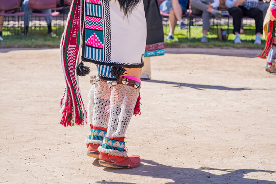Detailed view of colorful fringed skirt, beaded garters, and moccasins during a cultural performance on a dirt ground.