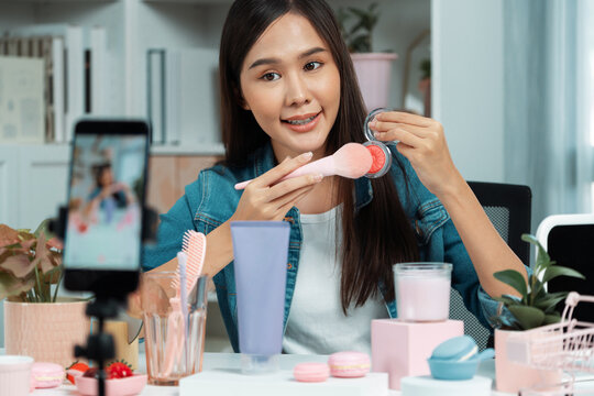 Young beautiful Asian reviewing cosmetic makeup brush cheek testing collection with pastel color, showing glow blusher mood with pink to promote on social media online recording smartphone. Stratagem.