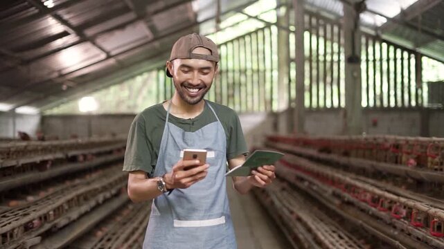 Near-end clip shows adult man centered in poultry house aisle, smiling while reviewing smartphone and notebook beside long rows of cages under soft daylight
