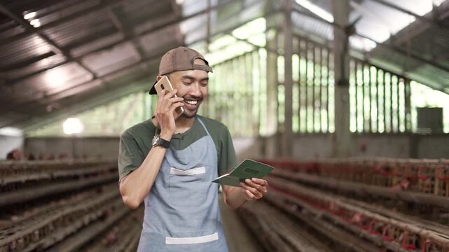 Throughout clip, farm worker stands inside poultry house, smiling while checking smartphone and clipboard, suggesting active farm coordination, record keeping