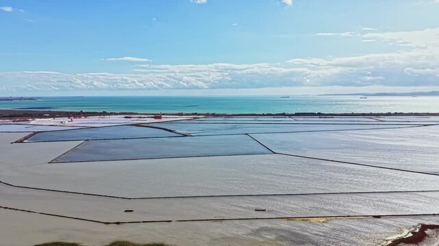 Lines of salt pans filled with water stretch across the landscape near the coast. The sky is blue with some clouds and the sea is visible beyond the pans.