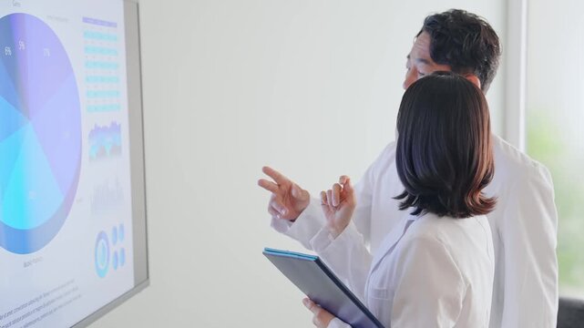 A group of men and women in white coats are talking while looking at a screen. Science, research, medicine, graphs, statistics, data, analysis, survey, testing.