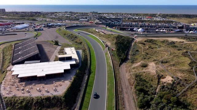 High-angle aerial drone view of Circuit Zandvoort race track curved section with moving sports car grandstands communications tower coastal dunes and North Sea horizon in North Holland Netherlands