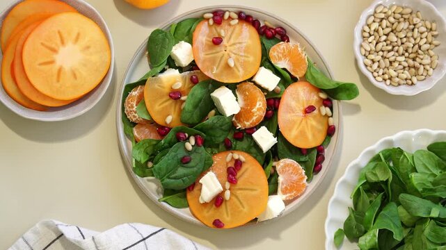 Overhead fresh spinach salad with sliced persimmon mandarin segments feta cubes pomegranate arils and pine nuts in bright natural light on neutral tabletop with bowls of ingredients nearby