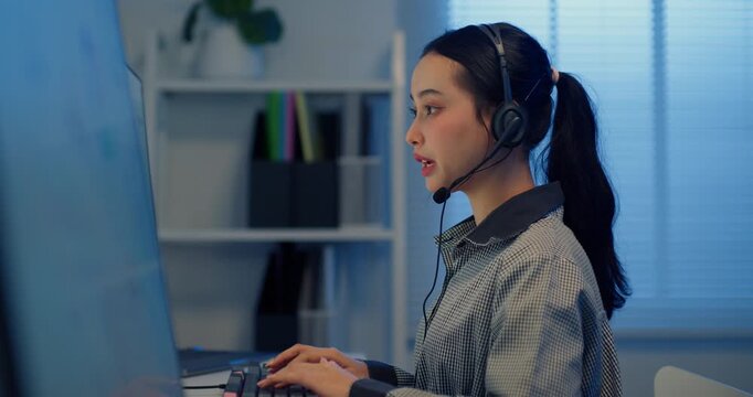 Side view of young Asian female call center agent wearing headset and talking while working on computer with business data dashboard in modern office.