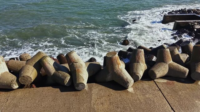 Concrete pieces are arranged along the edge of the water. Waves crash against the structures creating splashes. The sun shines down on the scene.