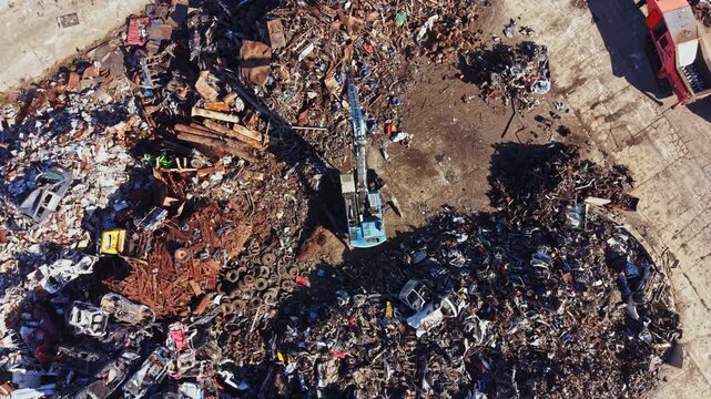 A large scrapyard is seen with piles of scrap metal and old vehicles. The area shows various parts of cars, metal scraps, and tires. The scene is during daylight.