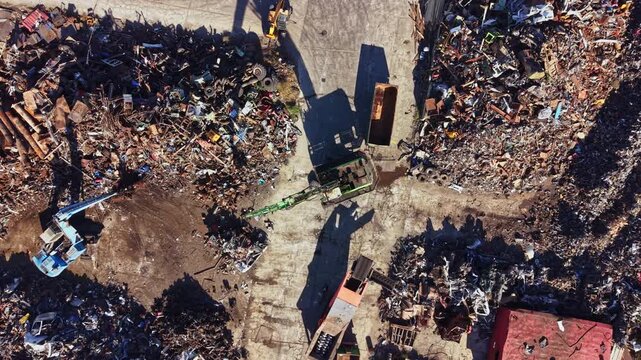 A large scrapyard is visible with piles of metal and machinery parts scattered throughout the area. Heavy equipment is used for sorting materials. The scene is bright and busy.