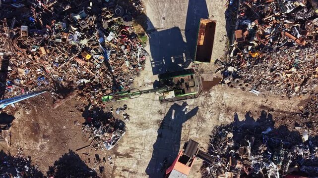 Machines are working in a scrap yard to sort and process various types of metal waste. The area is filled with piles of scrap materials under clear skies.