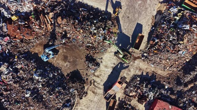 Aerial view of a yard filled with various metal scraps, discarded machinery, and debris. Workers are present among the piles, sorting materials for recycling.