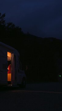 A Woman Steps Out Of A Camper Van At Night While Holding And Drinking Coffee From A Mug In An Outdoor Travel. Vertical, Static Shot