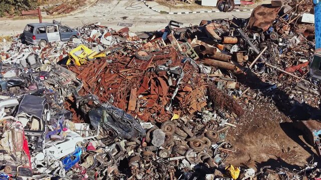 A collection of old cars and metal scraps are piled high in a yard. The scene shows a mix of rusted metal parts, tires, and vehicle remains. This location is near a busy road.