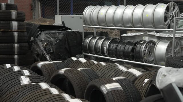 Rows of new car tires and bare alloy rims are stacked in an outdoor auto parts storage area on an overcast day.