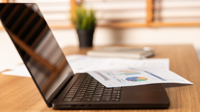 A close-up business scene an open laptop with a blank screen resting on a wooden desk. Business analysis reports with colorful charts and graphs are spread across the laptop and desk