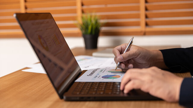 Professional business person analyzing financial reports with colorful charts and graphs on a laptop screen while working at a wooden desk in an office environment