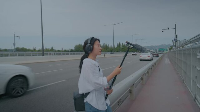 Young woman with headphones holding a boom microphone recording traffic sound on a city bridge road