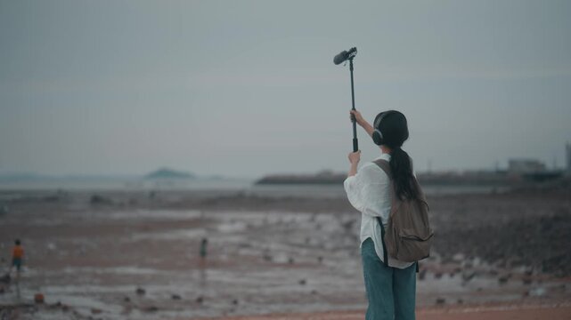 Female audio engineer wearing headphones and holding boom pole microphone on a coastal mudflat