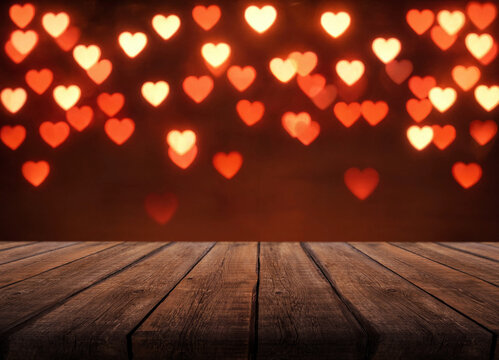 Heart shaped lights glow against dark red background above weathered wood table. Romantic bokeh forms warm backdrop, while broad uncluttered tabletop offers space for message or product display.