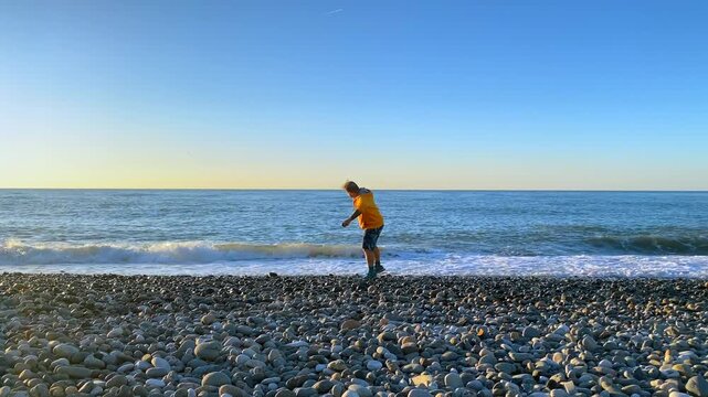 A man in a yellow sweatshirt stands on a pebble beach and throws stones into the Black Sea. The repeated motion and surf suit travel stories about seaside recreation in Batumi for coastal tourism.