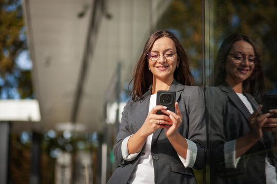 Smiling businesswoman checks smartphone beside reflective glass wall, keeping digital security in mind. Professional phone user reads her screen outdoors, with her reflection visible in window.