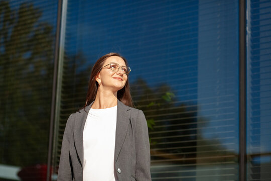 Thoughtful businesswoman looks toward sky in bright sunlight. Woman wearing eyeglasses and gray blazer stands before reflective corporate windows, smiling with calm, optimistic expression.
