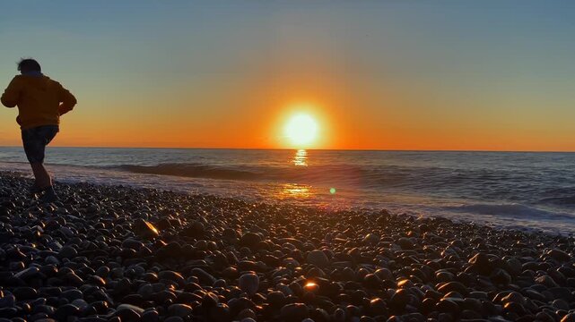 A man runs along a pebble beach at sunset as waves roll ashore. This coastal scene suits tourism campaigns, outdoor fitness promotions and travel content.