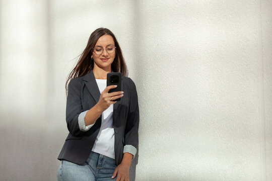 Professional woman smiles while reading smartphone screen outdoors beside bright textured wall. Glasses wearing businesswoman holds phone at chest height, dressed in dark blazer and jeans.