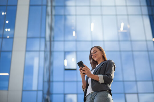 Businesswoman smiles while using smartphone beside glass office building. Professional in blazer reads her phone with both hands, taking relaxed pause in bright contemporary workplace setting.