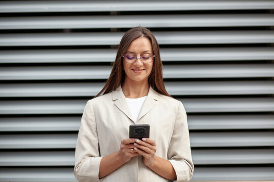 Woman uses smartphone with both hands and smiles softly, wearing eyeglasses and cream jacket. Office professional stands before striped metal wall, focused on screen with content expression.