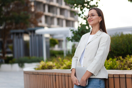 Confident woman poses with tablet beside wooden bench in landscaped office grounds. This smiling professional wears eyeglasses and pale blazer, with contemporary architecture and greenery behind her.