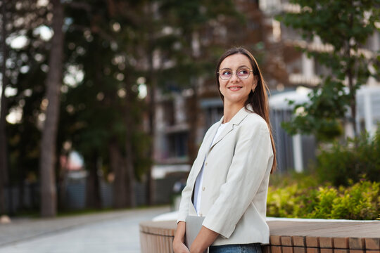 Confident woman stands beside wooden bench in landscaped city setting. Office professional in glasses and light blazer smiles gently, with greenery and buildings in soft focus, dressed in blazer